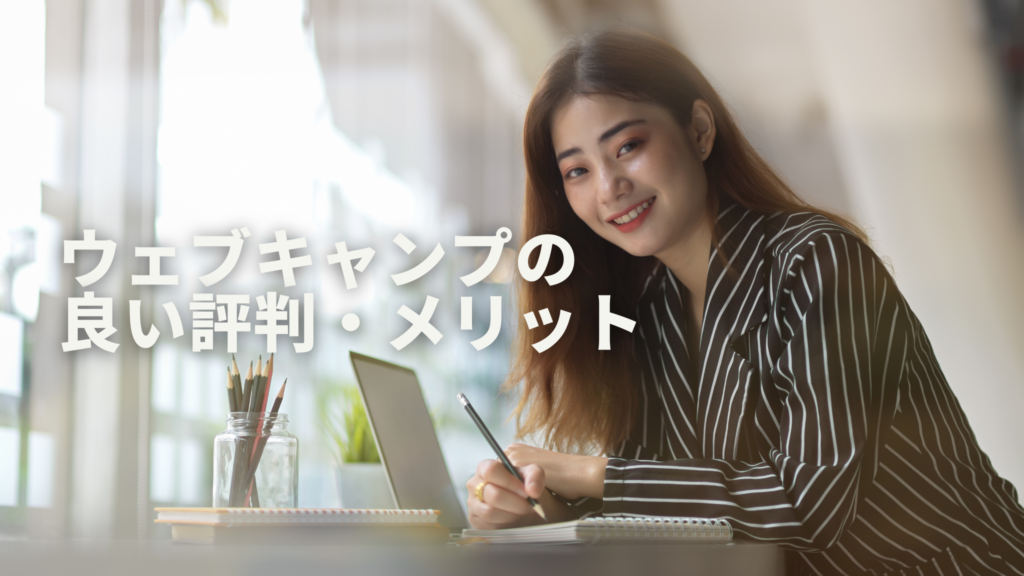 ロングヘアの女性がパソコンを開きながら鉛筆でノートにメモをとりながらこちらに笑顔を向けている
